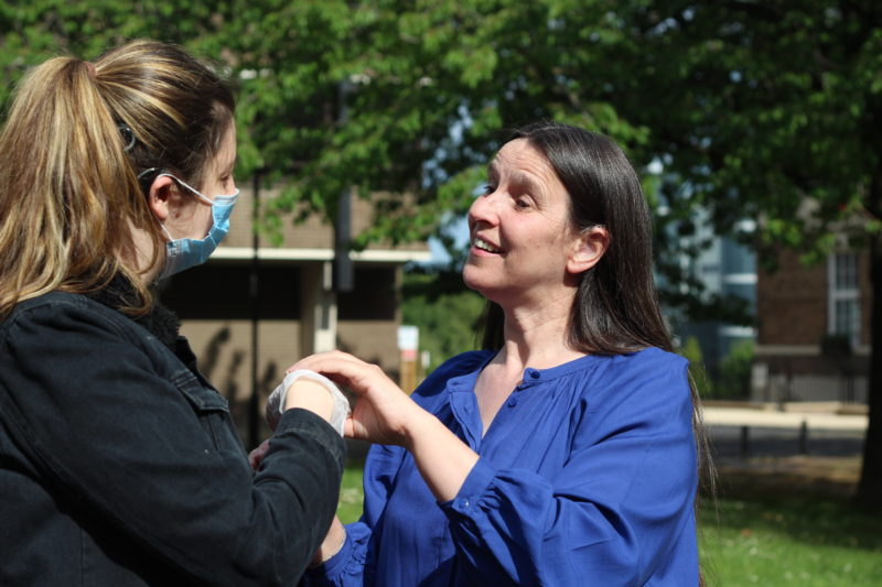 A woman wearing a mask and holding a hand of another deafblind woman.