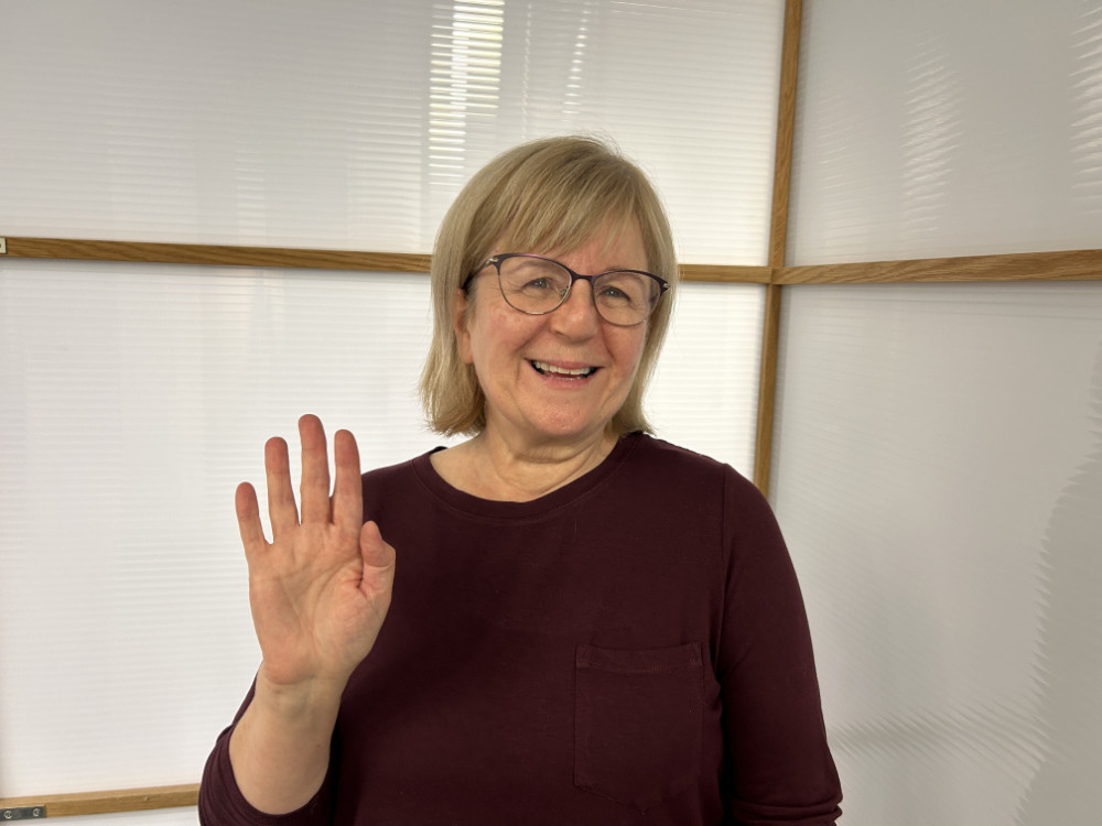 A woman in glasses is holding up a hand in front of a white wall.