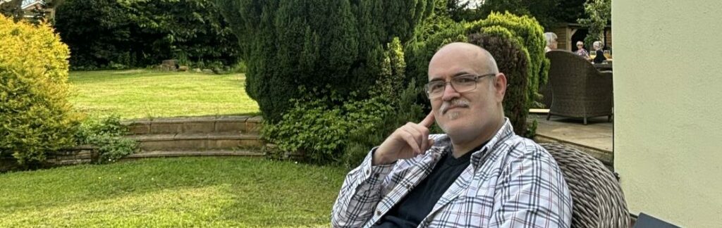 A man sits in a wicker chair outdoors, smiling slightly, with a lush green garden and trees in the background. He is wearing glasses, a plaid shirt, and dark pants.