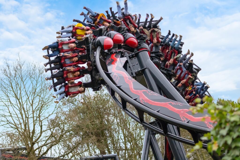 A group of people are riding a looping roller coaster, suspended upside down, against a backdrop of trees and a partly cloudy sky.