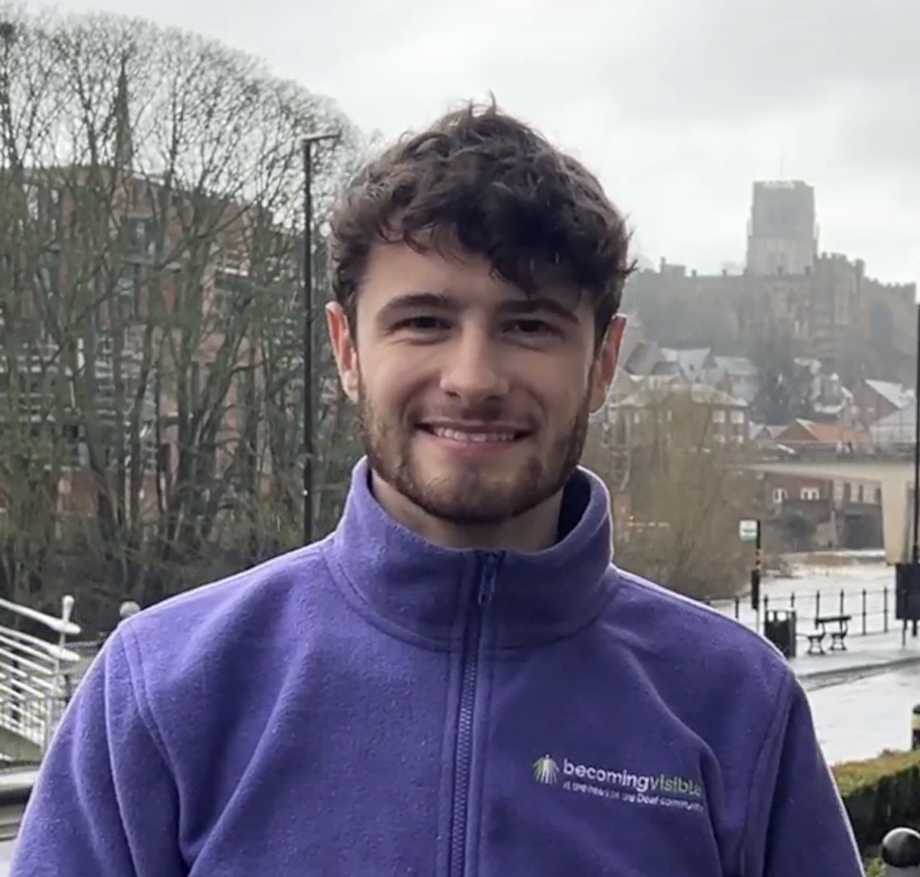 A man wearing a purple fleece jacket stands outdoors on a cloudy day with buildings, trees, and a distant church in the background.