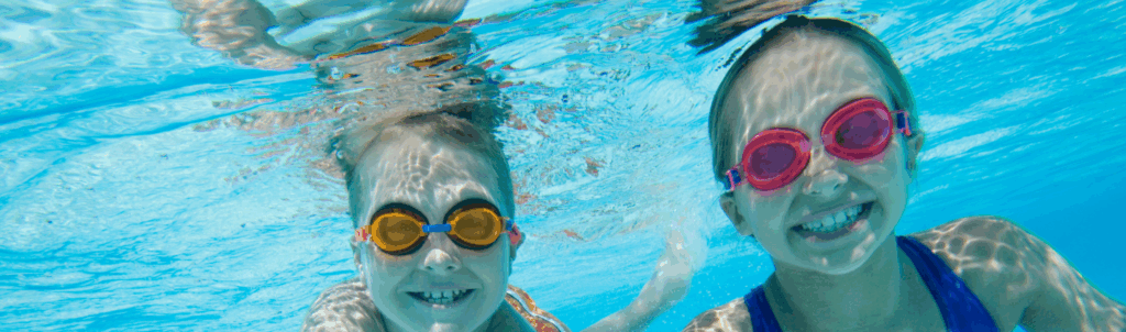 Two children wearing swim goggles smile and swim underwater in a pool.