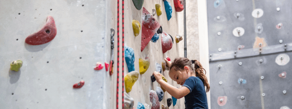 A young girl wearing a blue shirt climbs an indoor rock climbing wall with colorful holds.